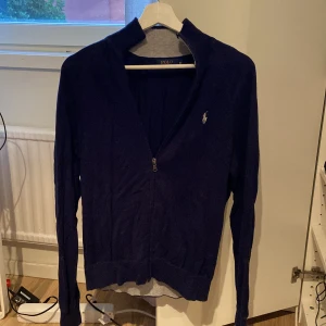 Marinblå kofta från Polo Ralph Lauren - Snygg marinblå kofta från Polo Ralph Lauren med dragkedja framtill och klassisk logga på bröstet. Tröjan har lång ärm, ribbade muddar och stårkrage. Perfekt för dig som gillar stilrena och tidlösa plagg.