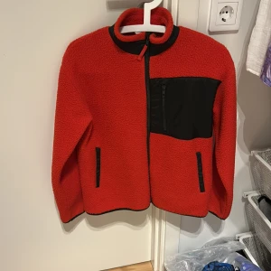 Röd teddyjacka med svarta detaljer - Röd teddyjacka med svarta detaljer och dragkedja. Jackan har en hög krage, svart bröstficka med dragkedja och två svarta fickor framtill. Perfekt för kalla dagar och riktigt skön att ha på sig.