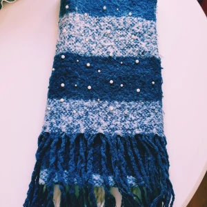 Blåvit randig halsduk med pärlor - Mysig och lång halsduk i blått och vitt med breda ränder och dekorativa pärlor. Halsduken är stickad i 100% akryl och har långa fransar i ändarna. Perfekt för att ge din outfit en cool touch under kalla dagar.  Helt & fint skick