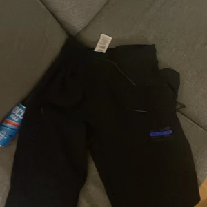 Svarta mjukisbyxor Emporio Armani M - Svarta mjukisbyxor från Emporio Armani med EA7-logga i silver och blått på benet. Byxorna har dragsko i midjan och en sportig look. Perfekta för chill eller träning, med bekväm passform och snygga detaljer.