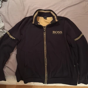 Svart track jacket från Hugo Boss - Svart track jacket från Hugo Boss med gulddetaljer och logga på bröstet. Jackan har hel dragkedja, ribbad krage med BOSS-tryck och guldfärgade ränder längs ärmarna. Perfekt för en clean och sportig look. Tröjan är nästan helt oanvänd och är i väldigt bra skick och är en storlek som passar alla