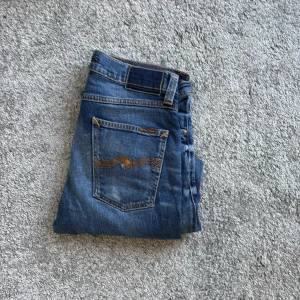 Blå jeans från Nudie Jeans Co, 29/34 - Säljer ett par klassiska blå jeans från Nudie Jeans Co i modellen 29/34. Jeansen har snygga kontrastsömmar, fem fickor och Nudie-loggan broderad på bakfickan. Tillverkade i slitstarkt bomullsjeans med en rak passform.