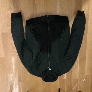 Quiltad bomberjacka från Zara Man - Mörkgrön quiltad bomberjacka från Zara Man med svarta muddar och dragkedja framtill. Jackan har två fickor med dragkedja och en klassisk bomberkrage. Perfekt för dig som gillar stilren och sportig look.