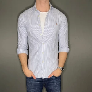 Randig skjorta från Ralph Lauren - Säljer en klassisk ljusblå och vit randig skjorta från Ralph Lauren med broderad logga på bröstet. Skjortan har lång ärm, knappar framtill och en snygg, normal passform. Perfekt till jeans eller chinos för en clean look.