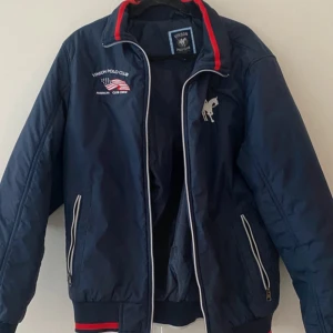 Blå vindjacka från Vinson Polo Club - Helt ny Snygg marinblå vindjacka från Vinson Polo Club med broderad logga och amerikansk flagga på bröstet. Jackan har vita dragkedjor, röda detaljer på kragen och två fickor med dragkedja. Perfekt för dig som gillar sportig och klassisk stil.