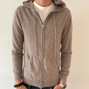 JOHN HENRIC KAHSMIR ZIPHOODIE - Mjuk och lyxig beige hoodie från John Henric i 100% kashmir. Tröjan har dragkedja, huva med snörning och två fickor framtill. Perfekt för dig som vill ha en stilren och bekväm look med premiumkänsla.