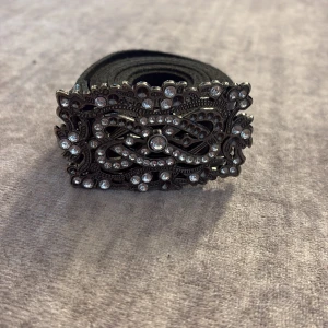 Svart bälte med glittrigt spänne - Snyggt svart bälte med ett stort, detaljerat spänne i antik silverfärg och massor av gnistrande stenar. Spännet har ett unikt, nästan vintage-inspirerat mönster som verkligen sticker ut. Perfekt accessoar för att lyfta vilken outfit som helst.
