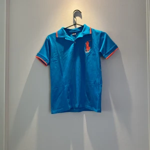 Blå pikétröja Ralph Lauren, strl 176 - Snygg blå pikétröja från Ralph Lauren med orange detaljer på krage och ärmslut. Stor orange logga på bröstet och broderad siffra 3 på ena ärmen. Klassisk krage och korta ärmar, perfekt för en sportig och stilren look.