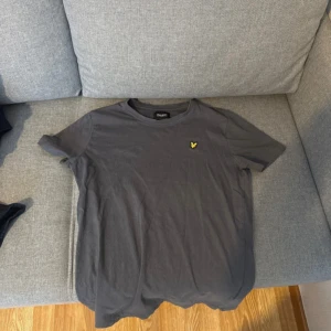 Mörkgrå t-shirt från Lyle & Scott - Snygg mörkgrå t-shirt från Lyle & Scott med klassisk rund hals och korta ärmar. På bröstet sitter den ikoniska gula örnloggan. Tillverkad i mjuk bomull som är skön mot huden. Perfekt för en clean och stilren look.