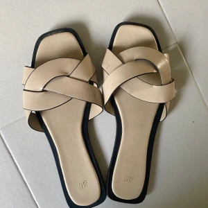 Beige sandaler med flätad rem H&M - Stilrena beige sandaler från H&M med flätad rem över foten och fyrkantig tå. Platt sula och svarta kanter ger en modern look. Perfekta för varma dagar och enkla att matcha med olika outfits.