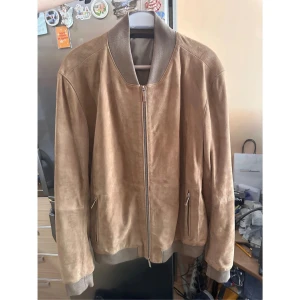 Beige mocka jacka Boss - Snygg beige bomberjacka i mocka med ribbade muddar och krage. Jackan har dragkedja framtill och två fickor med dragkedja. Perfekt för dig som vill ha en stilren och trendig look med en soft känsla. Inte använd alls NYPRIS:6499