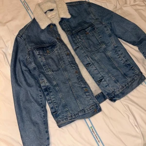 Jeansjacka med teddyfoder - Säljer en klassisk blå jeansjacka från logg med vitt teddyfoder och krage. Jackan har knappar framtill, två bröstfickor och långa ärmar. Perfekt för dig som vill ha en cool och tidlös look med extra värme.