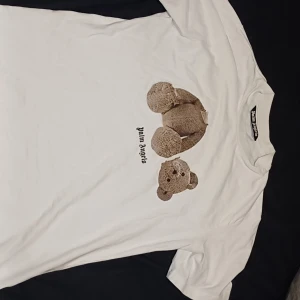 Vit Palm Angels t-shirt med nalletryck - Vit t-shirt från Palm Angels med ikoniskt nallebjörn-tryck på framsidan och stor svart logga på ryggen. Klassisk passform och rund halsringning. Materialet är mjuk bomull, perfekt för en avslappnad streetwear-look.