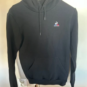 Svart hoodie från Le Coq Sportif - Svart hoodie från Le Coq Sportif med broderad logga på bröstet och fransk flaggdetalj på huvan. Klassisk känguruficka framtill, dragsko i huvan och långa ärmar. Mjuk bomullskänsla och relaxed fit – perfekt för chill dagar.