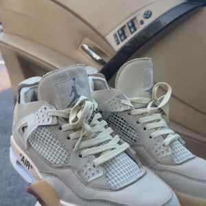 Nike Air Jordan 4 Off-White Sail - Exklusiva Nike Air Jordan 4 Off-White Sail med ikoniska detaljer som genomskinliga paneler, mesh och Off-White-typiska texten 'AIR' på sulan. Beige och vita toner, snörning och synlig Air-enhet i sulan. Perfekta för dig som vill sticka ut med en unik sneaker.