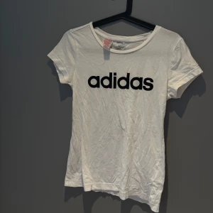 Vit t-shirt från Adidas med logga - Vit t-shirt från Adidas i bomull med klassisk svart logga tryckt framtill. Modellen är kortärmad med rund halsringning och har en clean, enkel stil som funkar till allt. Perfekt för dig som gillar sportiga och stilrena plagg. (Storleken är liten passar mig som har s/xs)