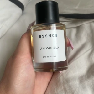 ESSNCE I Am Vanilla Eau de Parfum - Parfymflaska från ESSNCE med doften I Am Vanilla. Flaskan är rund och genomskinlig med svart lock och vit etikett. Innehållet är ljusgult och flaskan har en stilren, minimalistisk design.