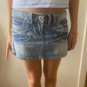 Blå jeanskjol från Awear - Säljer en snygg ljusblå jeanskjol från Awear med klassiska fickor fram och bak, bälteshällor och knappstängning. Kjolen är kort och har en avslappnad passform, perfekt till sommaren. Enkel att matcha med crop tops eller hoodies.