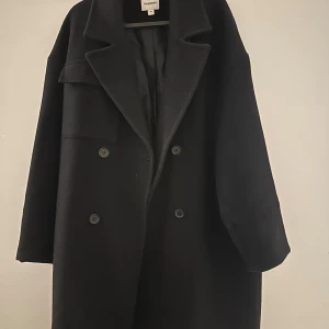 Mörk blå kappa från Kappahl - Stilren svart kappa från Kappahl med dubbelknäppning och bred krage. Oversized passform och långa ärmar ger en cool och avslappnad vibe. Perfekt för kalla dagar när du vill vara både varm och snygg.