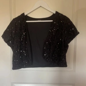Svart paljettprydd bolerotopp från Zara - Snygg svart bolerotopp från vila med glittrande paljetter. Perfekt för att lägga till lite glamour till din outfit. Kortärmad design som passar bra över en klänning eller topp.