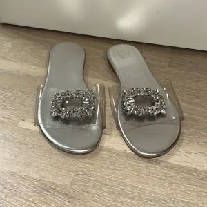 Silvriga sandaler med strassdetaljer - Snygga sandaler i silver med transparent rem och stora, glittriga strasspännen framtill. Platt sula och öppen tå ger en trendig look. Perfekta för dig som vill sticka ut med lite extra bling.