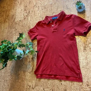 Röd pikétröja från Polo Ralph Lauren - Klassisk röd pikétröja från Polo Ralph Lauren med blå broderad logga på bröstet. Tröjan har korta ärmar, krage och två knappar framtill. Tillverkad i bomull och har en normal passform. Perfekt för en clean och stilren look. Den är Slim fit i XS/XXS så om man vill ha dendär looken när den sitter tajt så är detta en perfekt tröja för dig som har storlek XXS