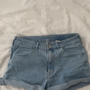 Ljusblå jeansshorts från &Denim - Snygga ljusblå jeansshorts från H&M i stretchigt material. Modellen har regular waist, fickor och uppvikta benslut. Perfekta för varma dagar och enkel att matcha med allt. Stängs med dragkedja och knapp framtill.