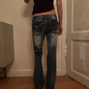 Blå bootcut jeans med slitningar - Säljer ett par blå bootcut jeans med coola slitningar och detaljerade bakfickor. Jeansen har låg midja och en tvättad look som ger en avslappnad vibe. Perfekta för dig som gillar en lite baggy och loose passform. Innerbenslängden är 78cm och midjan är 38-39 cm