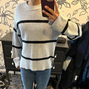 Stickad tröja - Säljer en ribbstickad tröja i ljusgrått med svarta horisontella ränder. Tröjan har en avslappnad passform, rund halsringning och långa ärmar med breda muddar. Perfekt för dig som gillar en chill och stilren look.