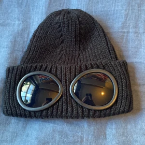 Svart C.P goggle-mössa - Cool svart stickad mössa med ikoniska goggle-linser framtill. Mössan är ribbstickad och har en uppvikt kant där de runda glasögon-liknande detaljerna sitter. Perfekt för dig som vill sticka ut med streetwear-vibe.