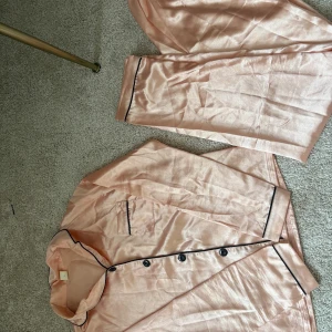 Champagnefärgad pyjamas i satin - Snygg pyjamas i champagnefärgad satin med svarta detaljer. Setet består av långärmad skjorta med knappar och bröstficka samt matchande byxor. Mjukt och glansigt material som känns lyxigt mot huden. Perfekt för chillkvällar hemma. Storlek 146/152. Liten fläck på ärmen som man knappt tänker på därav priset.💓