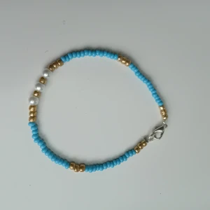 Blått pärlarmband med gulddetaljer - Handgjort armband skapat med små blå pärlor, guldfärgade pärlor och några vita pärlor. Armbandet har ett silvrigt lås och är perfekt för att ge din outfit en färgklick. Enkel och trendig design som passar till många stilar.