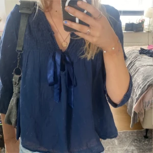 Odd Molly - Supersöt mörkblå blus med smockad överdel och v-ringning. Blusen har ett fint knytband framtill och långa ärmar med resår vid handlederna. Luftig och lätt i materialet, perfekt till jeans eller kjol.
