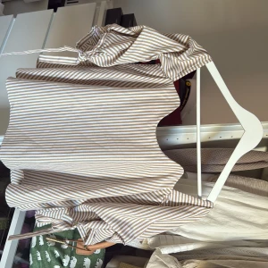 Randig kortklänning från SM Collection - Superfin topp från SM Collection med beige och vita ränder. Toppen har en söt volangkant nertill, dragkedja i sidan och knytband vid halsen. Perfekt för sommarens alla tillfällen och ger en fräsch och trendig look.