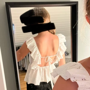 Vit volangtopp med öppen rygg - Supersöt vit topp med volangärmar och broderade detaljer. Toppen har fyrkantig ringning fram, öppen rygg med knytband och en lätt utsvängd peplum-nederdel. Perfekt för sommarens alla tillfällen och ger en romantisk vibe.