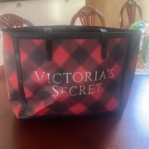 Rutig shoppingväska Victoria's Secret - Stor och rymlig shoppingväska från Victoria's Secret med röd och svart rutig design. Väskan har svarta handtag och tydlig logga framtill. Perfekt för att bära med sig allt man behöver under dagen. Aldrig använd och ser ut som ny 