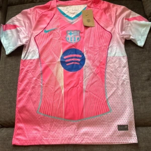 FC Barcelona x Nike rosa fotbollströja - Snygg FC Barcelona fotbollströja från Nike i rosa och vita toner med turkosa detaljer. Tröjan har korta ärmar, V-ringning och är tillverkad i lätt, ventilerande Dri-FIT-material. Framsidan pryds av klubbmärke och sponsorlogga i blått. Perfekt för dig som vill sticka ut på planen.
