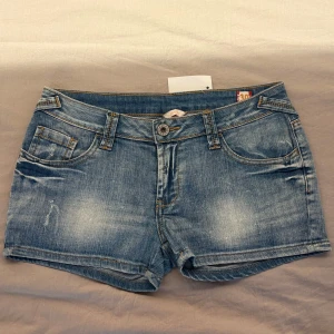 Blå lågmidjade jeansshorts  - Snygga blå lågmidjade jeansshorts med klassisk femficksdesign, slitna detaljer och röda sömmar vid bakfickorna. Shortsen har normal passform, bälteshällor och knappstängning framtill. 