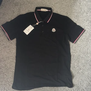 Svart Moncler pikétröja med röd/vit kant - Svart pikétröja från Moncler med klassisk krage och knäppning framtill. Röd och vit rand längs krage och ärmslut ger en clean kontrast. Broderad Moncler-logga på bröstet. Tillverkad i mjuk bomull för skön passform.