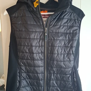 Svart dunväst Parajumpers Warm Up L - Snygg svart dunväst från Parajumpers i storlek L. Barn strlk motsvarar en xs small.Västen har quiltad framsida, huva och coola detaljer som gul knapp och orange hängare. Materialet är polyester och syntet, perfekt för lager-på-lager. Dragkedja framtill och broderad logga på axeln. Nypris 3900:-