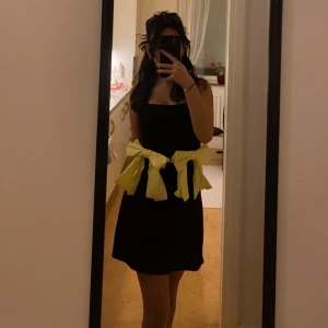 Svart kort klänning med gula rosetter - En svart, ärmlös kort klänning med raka axelband och två stora gula rosetter i midjan. Klänningen har en enkel och stilren design med en färgglad twist tack vare rosetterna. Perfekt för dig som vill sticka ut med en cool detalj.