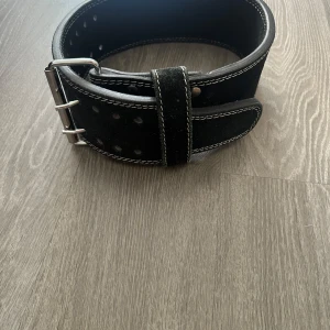 Svart lyftarbälte Gorilla Wear - Säljer ett svart lyftarbälte från Gorilla Wear med vit kontrastsöm och bred design för extra stöd. Bältet är gjort i mocka och har ett stort logomärke framtill. Perfekt för dig som vill maxa på gymmet och lyfta tungt.