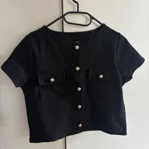 Svart kortärmad topp från H&M Divided - Stilren svart topp från H&M Divided med korta ärmar och croppad passform. Toppen har två fickor framtill med dekorativa knappar i silverfärg och knäppning hela vägen fram. Perfekt för en trendig och clean look. Spontan köp från ca 6/7 månader sen!! Aldrig använt! Original pris: 249kr säljs för 100kr! Har bara legat i garderoben sen jag köpte den!😅..