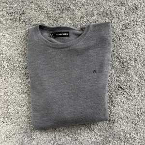Grå tröja från J.Lindeberg - Snygg grå tröja från J.Lindeberg med minimalistisk design och liten broderad logga på bröstet. Tröjan har rund halsringning och är tillverkad i mjuk merinoull, perfekt för en clean och stilren look. Obs tagen håller på att ramla av sitter bara fast på ena sidan.
