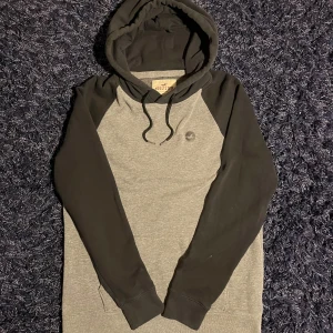 Hollister hoodie - Snygg hoodie från Hollister i grått och svart med klassisk logga på bröstet. Tröjan har svarta ärmar och huva med snörning, samt känguruficka framtill. Perfekt för chill dagar eller när du vill ha en avslappnad stil. Hör av er vid minsta fundering🙌
