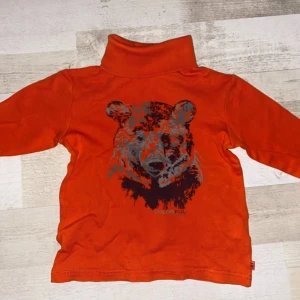 Orange polotröja med björntryck - Snygg orange polotröja med lång ärm och ett stort tryck av ett björnhuvud i svart och grått framtill. 