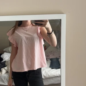 Ljusrosa volangblus från Gina Tricot - Supersöt ljusrosa blus från Gina Tricot med volangdetaljer på ärmarna. Blusen är ärmlös och har en loose passform. Tröjan är från gina tricots barnavdelning i storlek 170