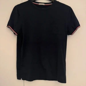 Svart Moncler t-shirt med logga - Svart t-shirt från Moncler med klassisk passform, rund hals och korta ärmar. Ärmsluten har snygga ränder i vitt och rött samt en broderad Moncler-logga på ena sidan.Perfekt för en clean och stilren look.