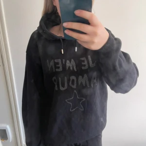 Svart hoodie med text och stjärna - Svart hoodie med glittrig texten 'JE M'EN AMOUR' framtill och en stjärna under. Tröjan har huva med snörning och långa ärmar. Pris kan diskuteras. Storlek 158/164 men passar xs/s