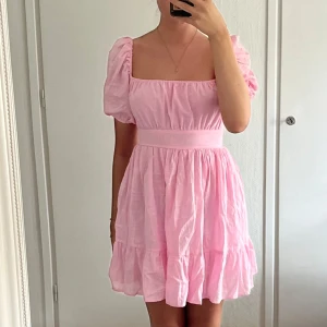 Söt rosa kortklänning med puffärm - Superfin ljusrosa kortklänning med puffärmar, fyrkantig urringning och volangkant nertill. Klänningen har öppen rygg med stor rosett och dragkedja i midjan. Perfekt för sommarens alla tillfällen!
